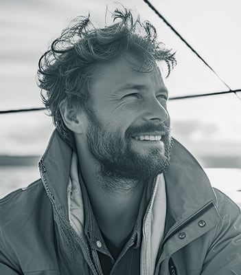 Mann mit Bart und lockigen Haaren trägt eine Jacke und lächelt, während er auf einem Boot sitzt.