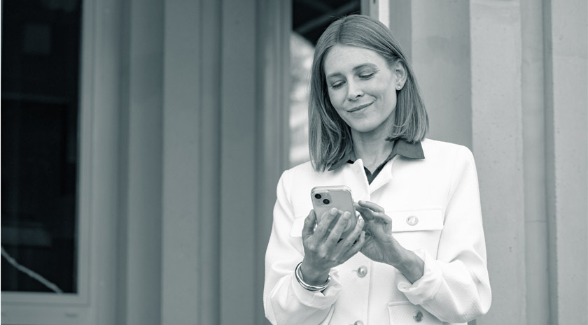 Eine Frau mit schulterlsngen, hellen Haaren und einem schicken Mantel blickt zufrieden auf ihr Smartphone.