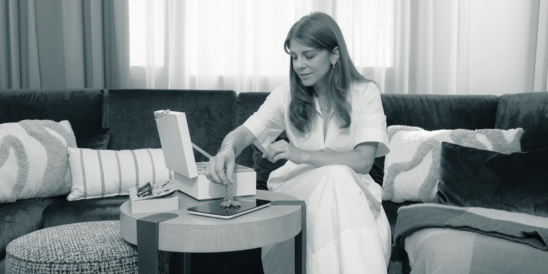 Eine junge Frau sitzt in einem schicken Kleid auf einer modernen Couch und wiegt Goldschmuck, den sie aus einem Schmuckkästchen holt.