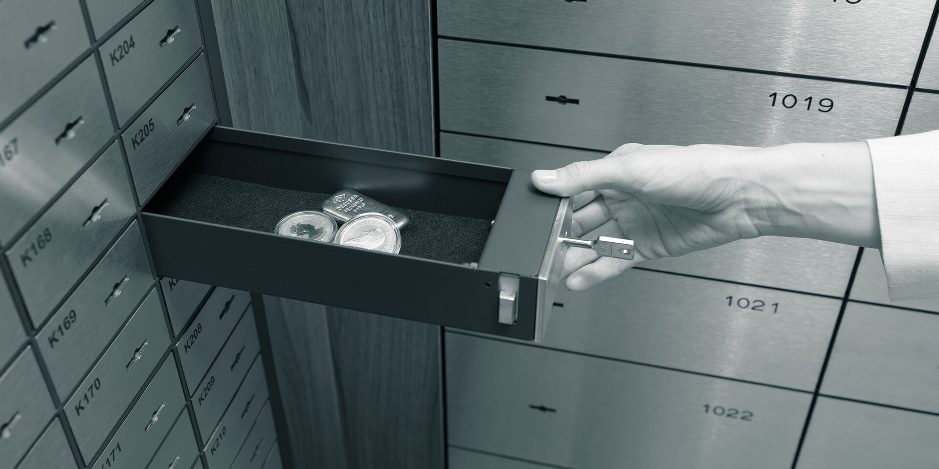 Eine Hand öffnet eine Schließfach-Schublade in einem Raum voller Schließfächer. Darin liegen Münzen und ein Goldbarren.