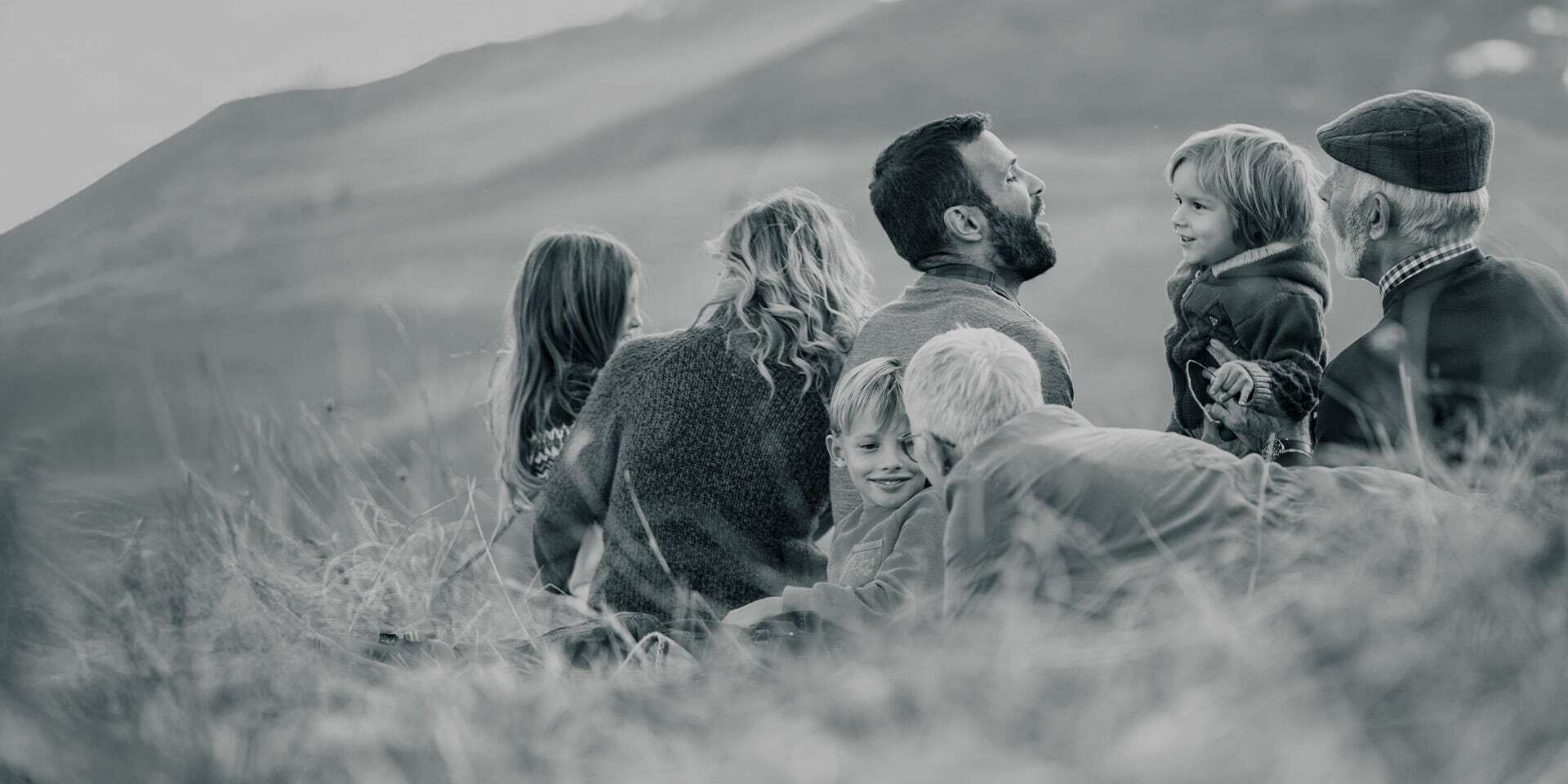 Mehrgenerationen-Familie sitzt gemeinsam im Gras und unterhält sich lachend.
