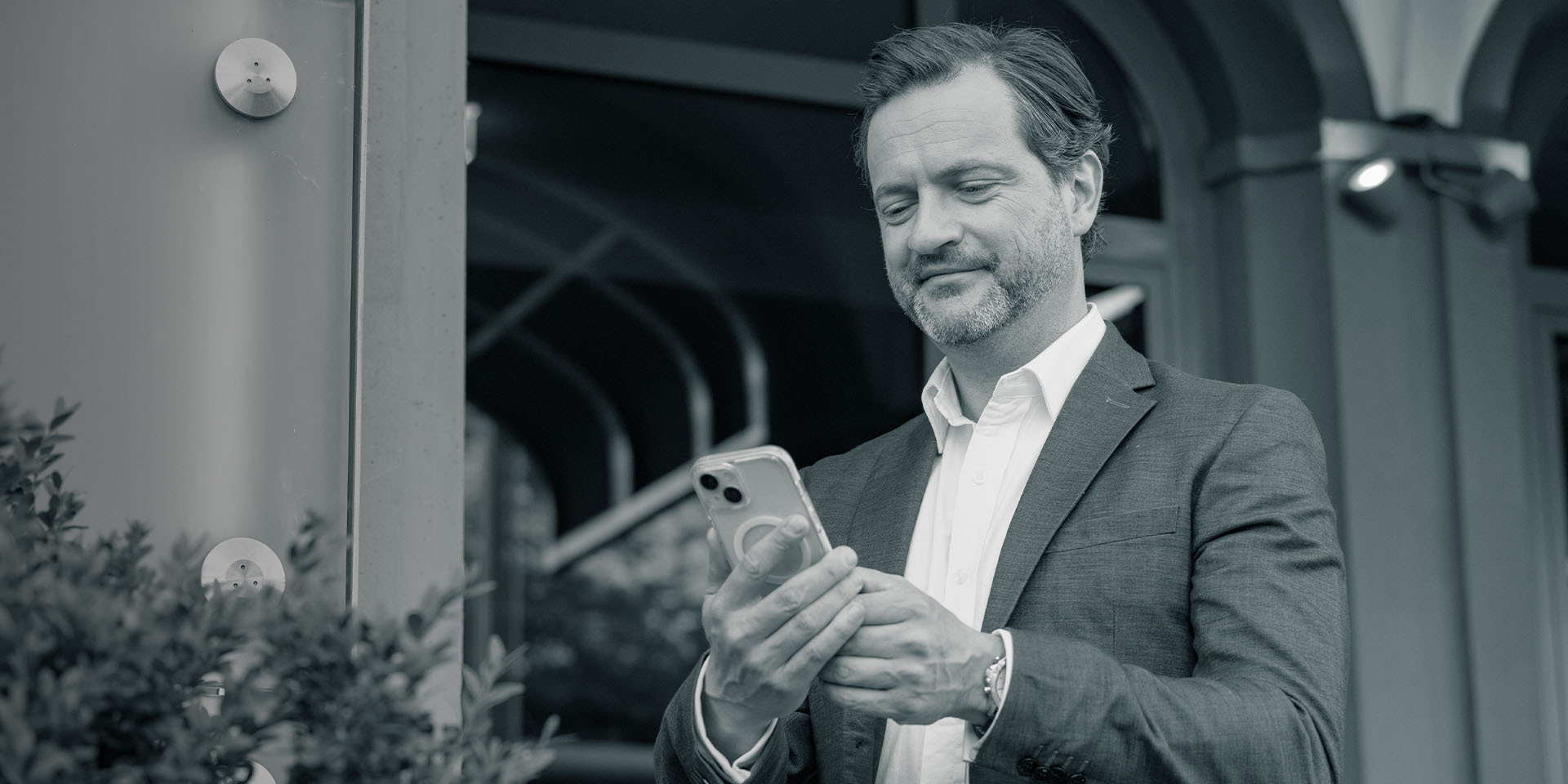 Geschäftsmann im Anzug steht vor einem Gebäude und blickt zufrieden auf sein Smartphone.