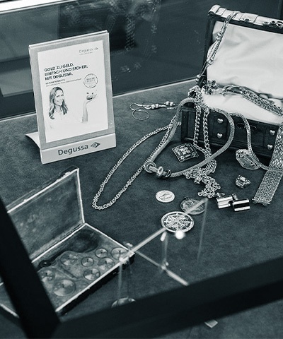 Detailaufnahme einer Auslage in einer Glasvitrine mit Gold- und Silberschmuck, Münzen, Manschettenknöpfen und einer Truhe, sowie einer Werbeaufsteller