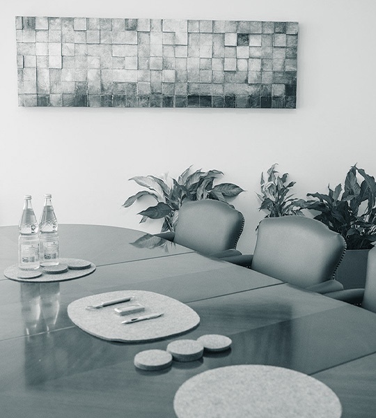 Konferenztisch aus dunklem Holz mit Filzunterlagen, Wasserflaschen und einem rechteckigen Kunstwerk an der Wand im Hintergrund