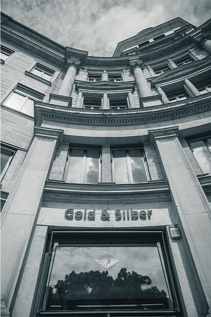 Aufwärtsansicht der gewölbten Fassade der Degussa Niederlassung in München, mit Säulen, Fenstern und dem Schriftzug \"Gold & Silber\" über dem Schaufenster