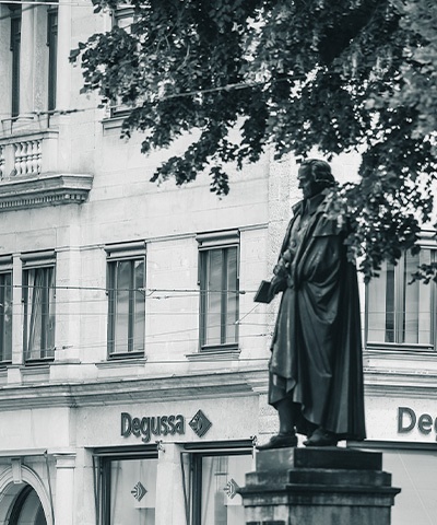 Aufnahme einer Statue auf einem Sockel, teilweise verdeckt von einem Baum, vor der Fassade der Degussa Niederlassung München