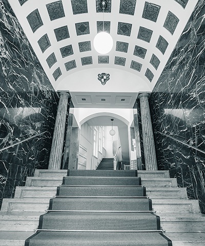 Elegantes Treppenhaus zum Eingang der Nürnberger Niederlassung mit dunklen Marmorwänden, verzierten Deckenkassetten, zwei Säulen und einem zentralen Teppich auf den Stufen