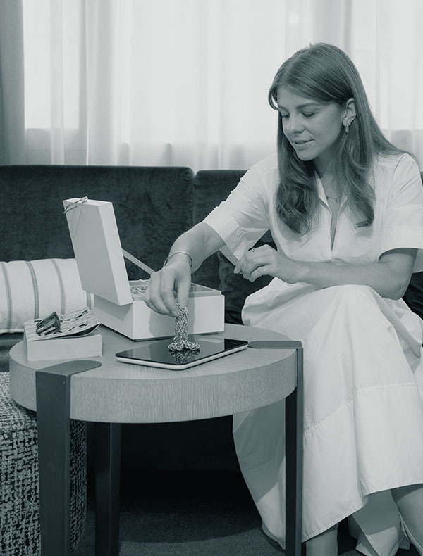 Frau in weißem Kleid sitzt im Wohnzimmer und legt eine Goldkette auf eine Waage, neben ihr eine geöffnete Schmuckschatulle.