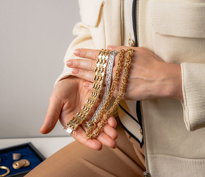 Person hält mehrere goldene und silberne Armketten in der Hand, im Hintergrund liegt weiterer Schmuck in einem Etui.