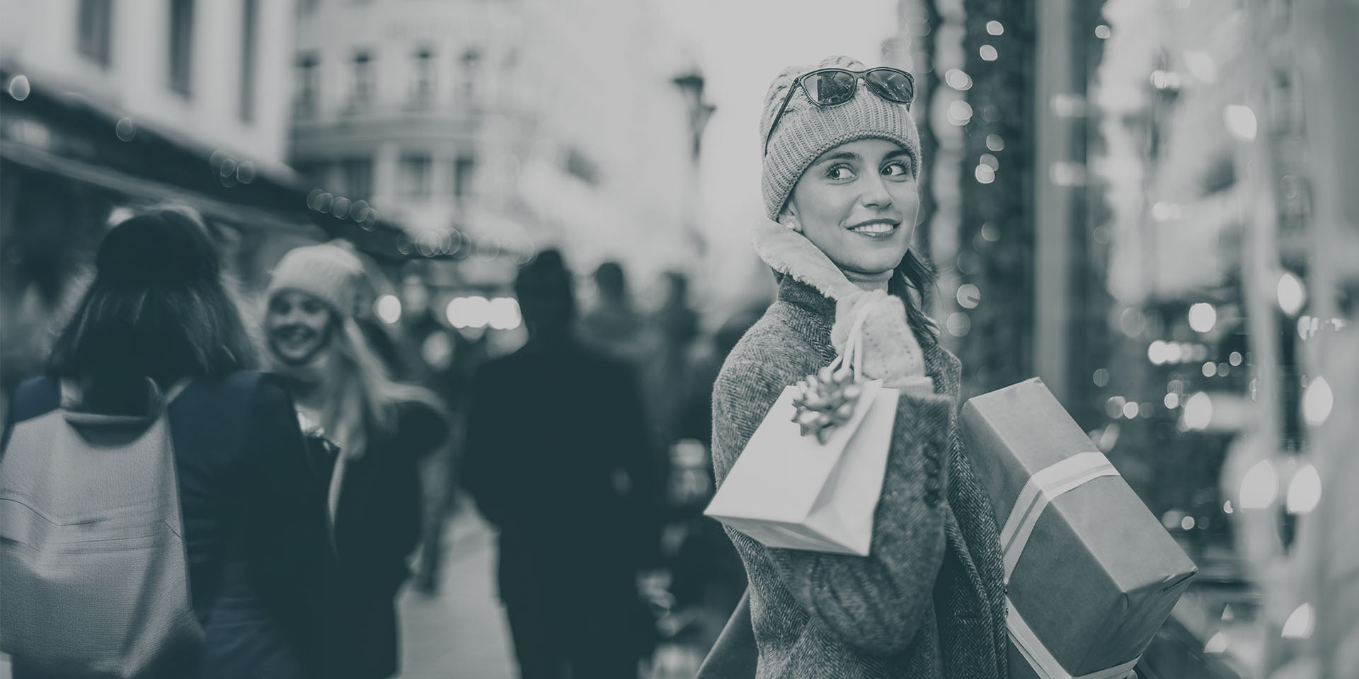 Frau mit Mütze und Winterkleidung trägt mehrere Geschenktüten und ein verpacktes Paket, während sie lächelnd durch eine weihnachtlich beleuchtete Einkaufsstraße geht