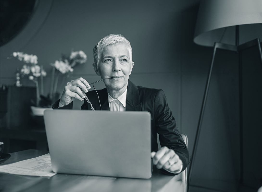 Geschäftsfrau mit kurzen grauen Haaren sitzt am Laptop und hält nachdenklich ihre Brille in der Hand