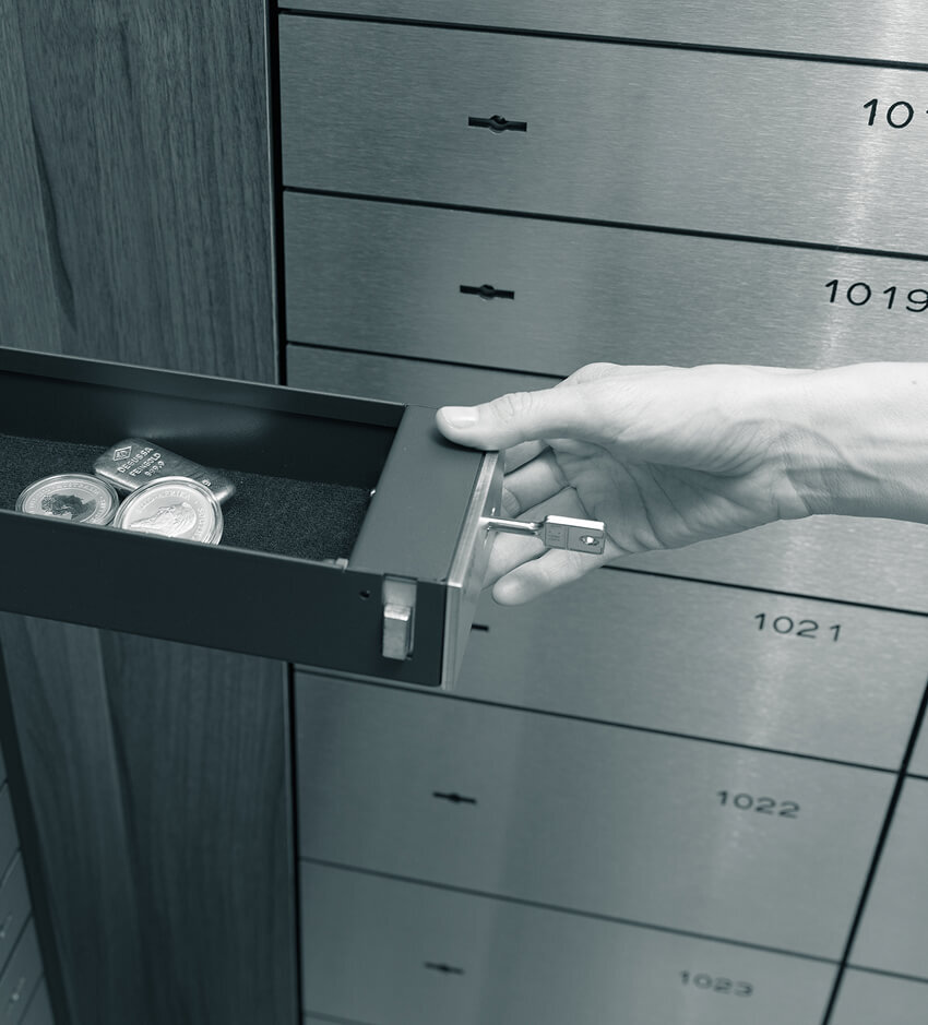 Eine Hand zieht ein geöffnetes Schließfach aus derm Schrank. Darin befinden sich Goldmünzen und -barren