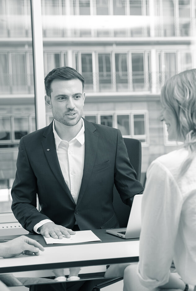 Ein junger, männlicher Degussa-Mitarbeiter im Anzug berät eine Kundin, die ihm Gegenüber am Schreibtisch sitzt.